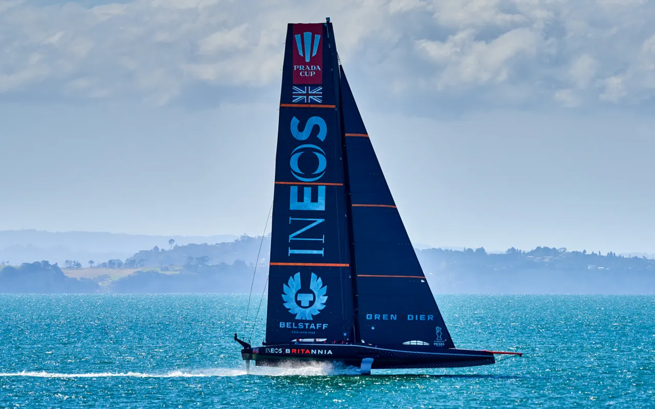 A dark-hulled AC75 foiling yacht with a tall black mainsail