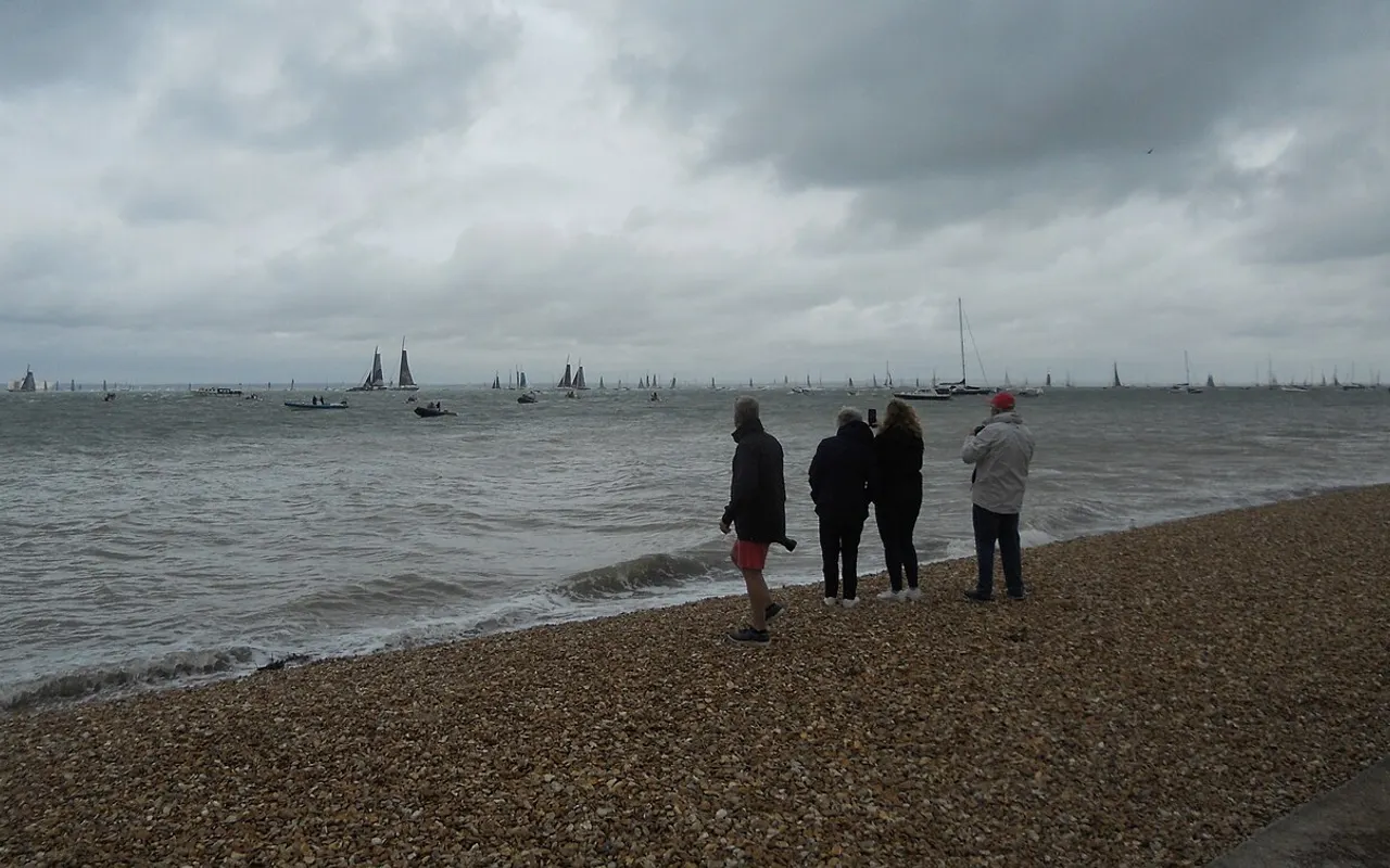 Spectators on a shingle beach watching yachts start the Fastnet Race off Cowes