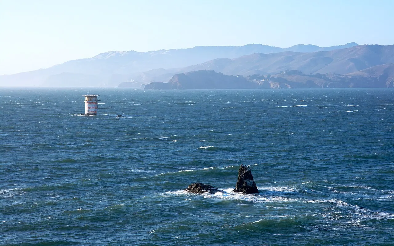 Un petit phare sur des rochers au large d'une côte vallonnée, avec du ressac sur les rochers voisins