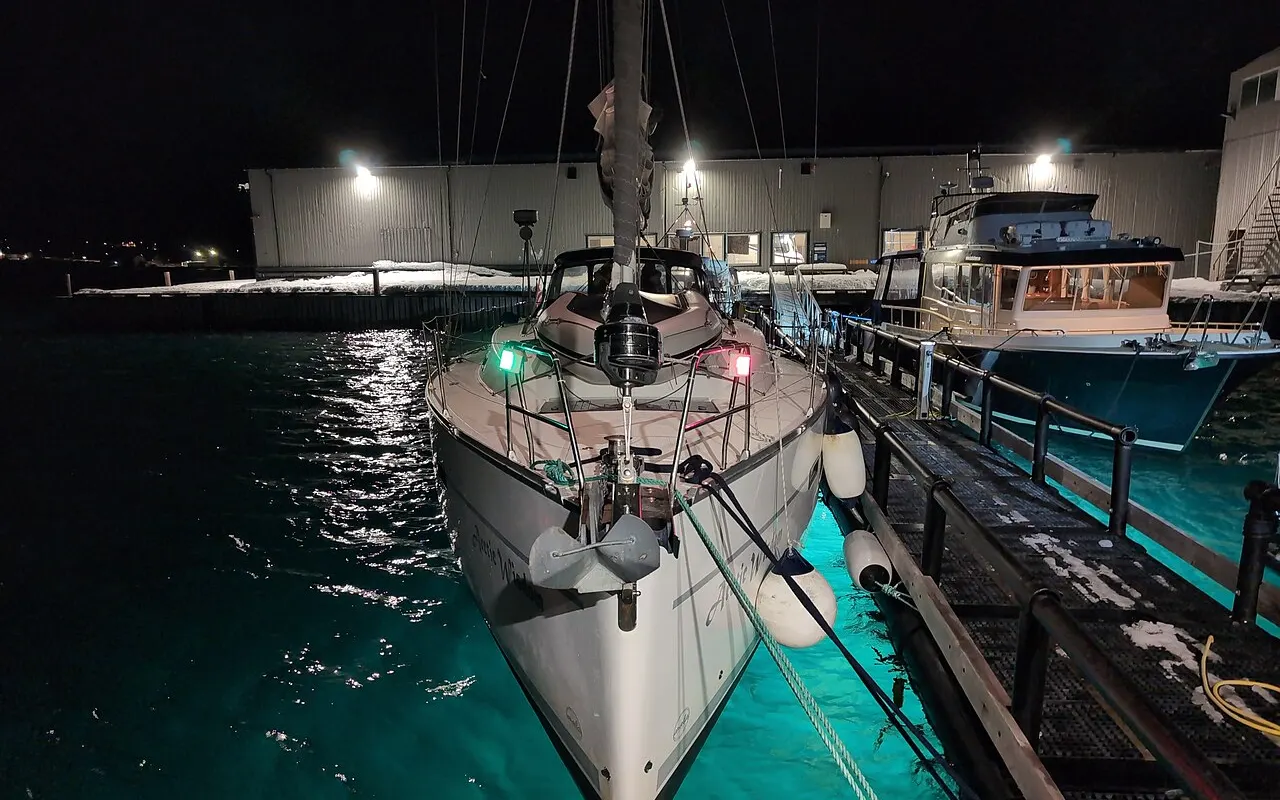 A sailing yacht's bow at night in a marina, its red and green sidelights lit