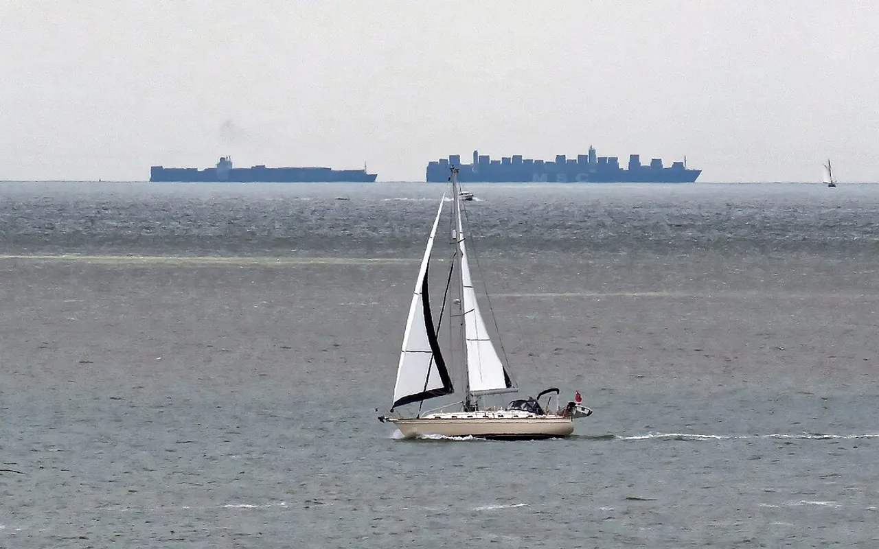 A sailing yacht under way with large ships on the horizon