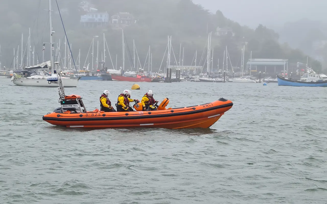 An orange RNLI inshore lifeboat with crew in a misty harbour at Dartmouth