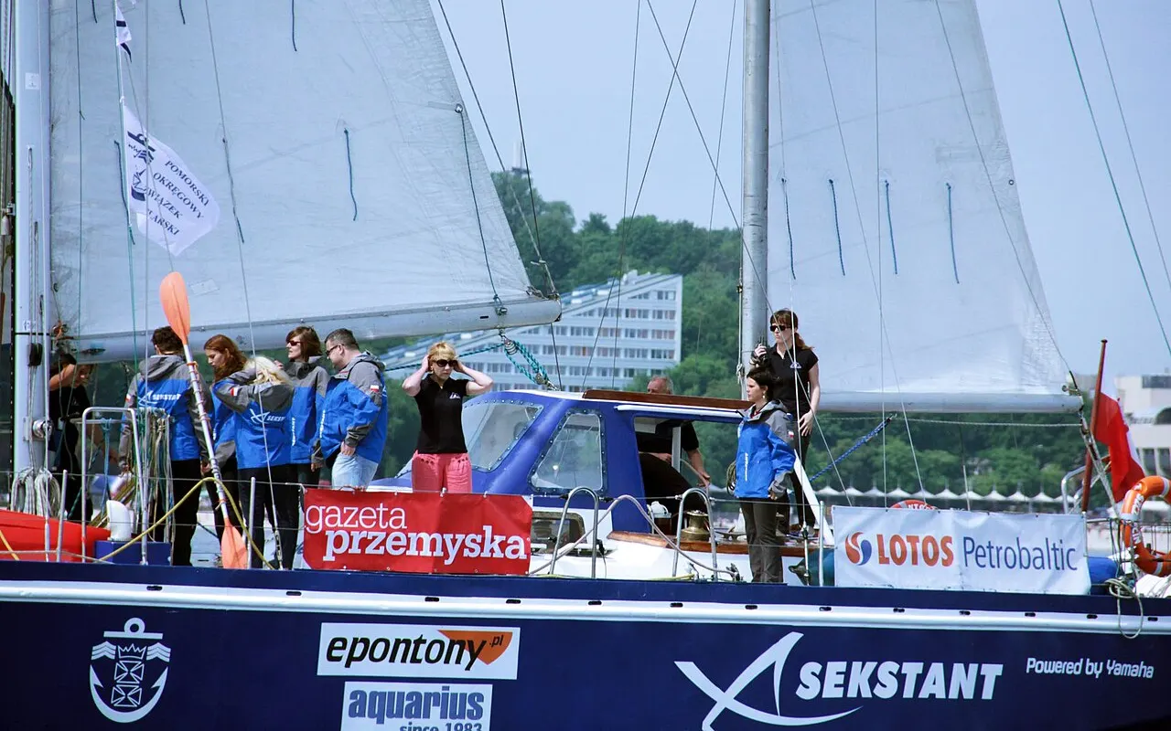 A sail training crew in matching jackets lining the deck of a yacht at a harbour