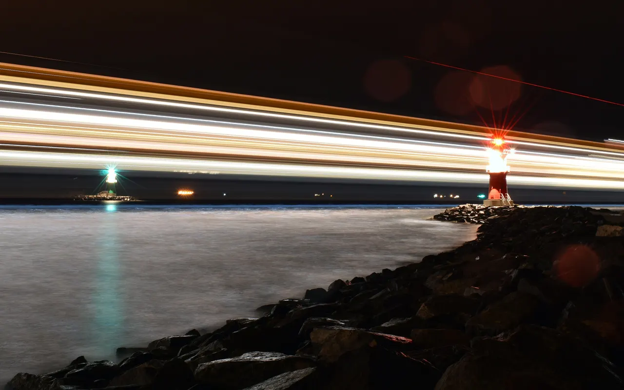 Long exposure at night of a ship's lights streaking past a harbour light on a breakwater