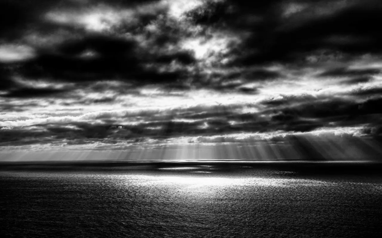 Black and white photo of a dark sea under heavy clouds with sunlight breaking through