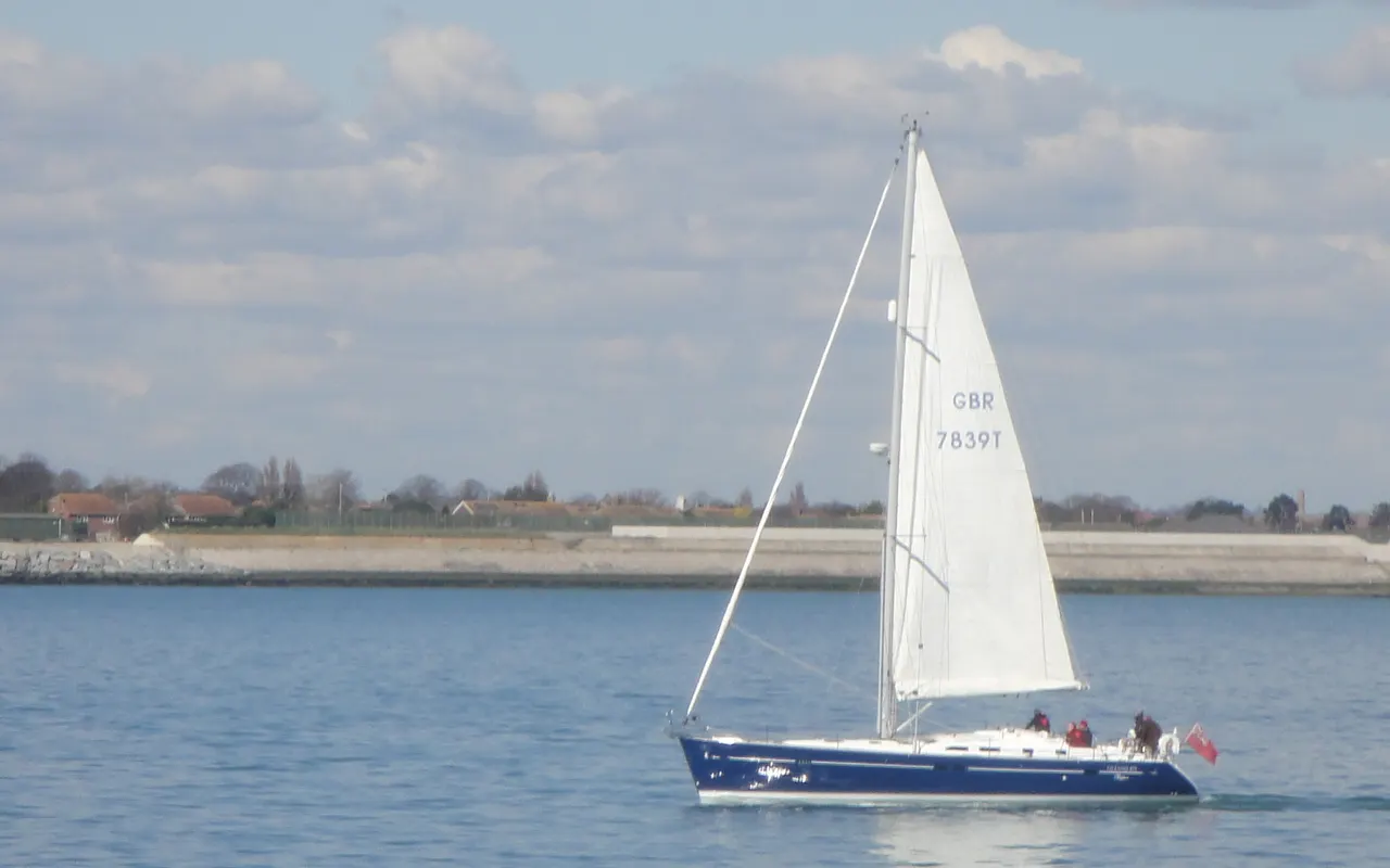 A white sloop sailing in the Solent with the shore behind
