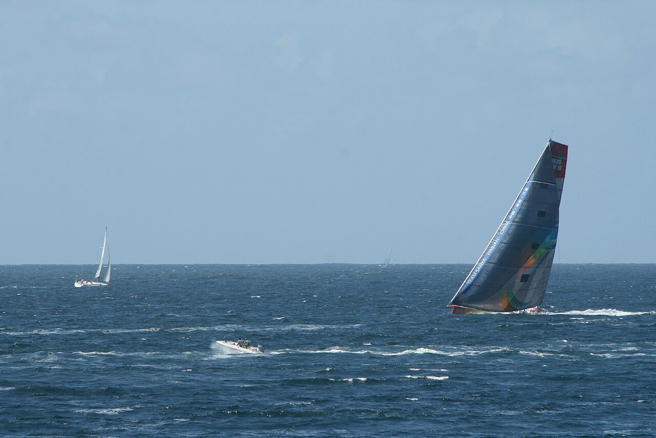 A Volvo Open 70 racing yacht heeled hard under sail, with a cruising yacht in the background